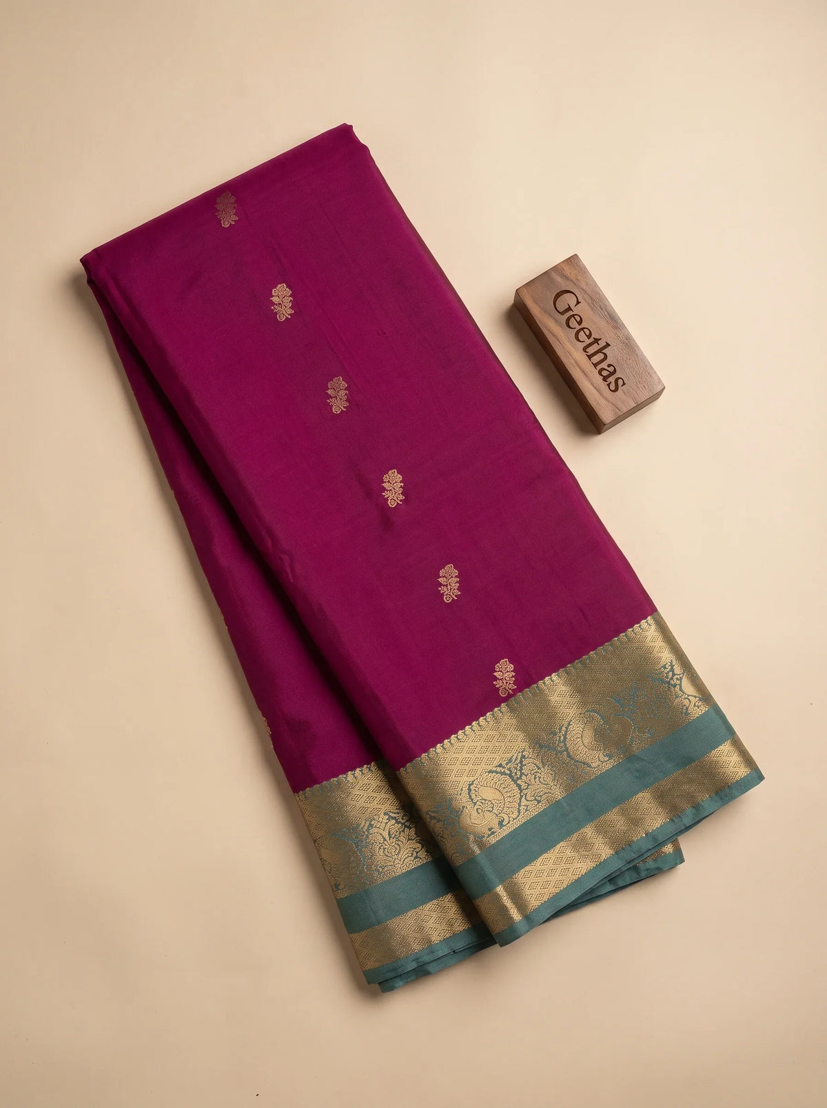 Folded magenta soft silk saree with small gold-toned floral motifs and teal decorative woven border