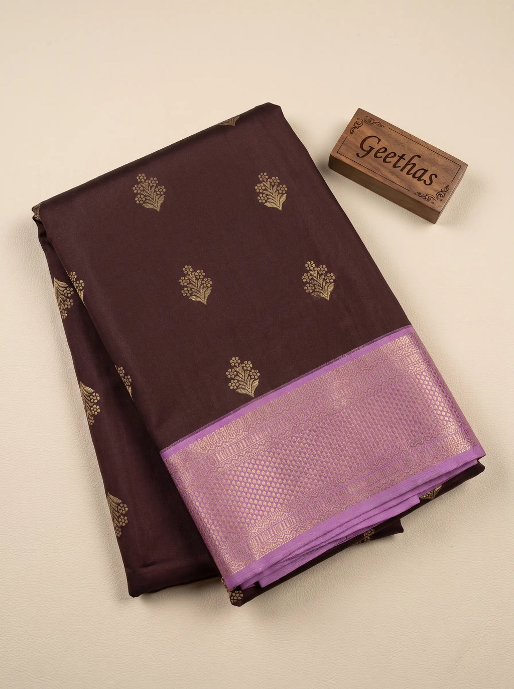 Folded deep brown soft silk saree with gold-toned woven motifs and lilac geometric border