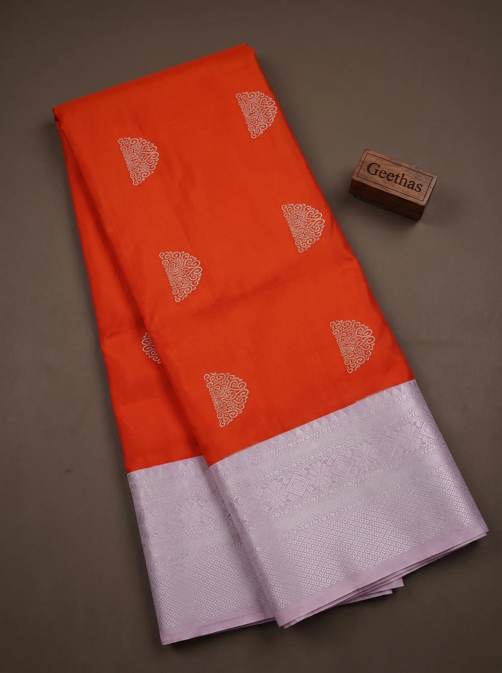 Vermilion orange soft silk saree displayed flat with woven paisley-inspired motifs and a lavender-toned patterned border.